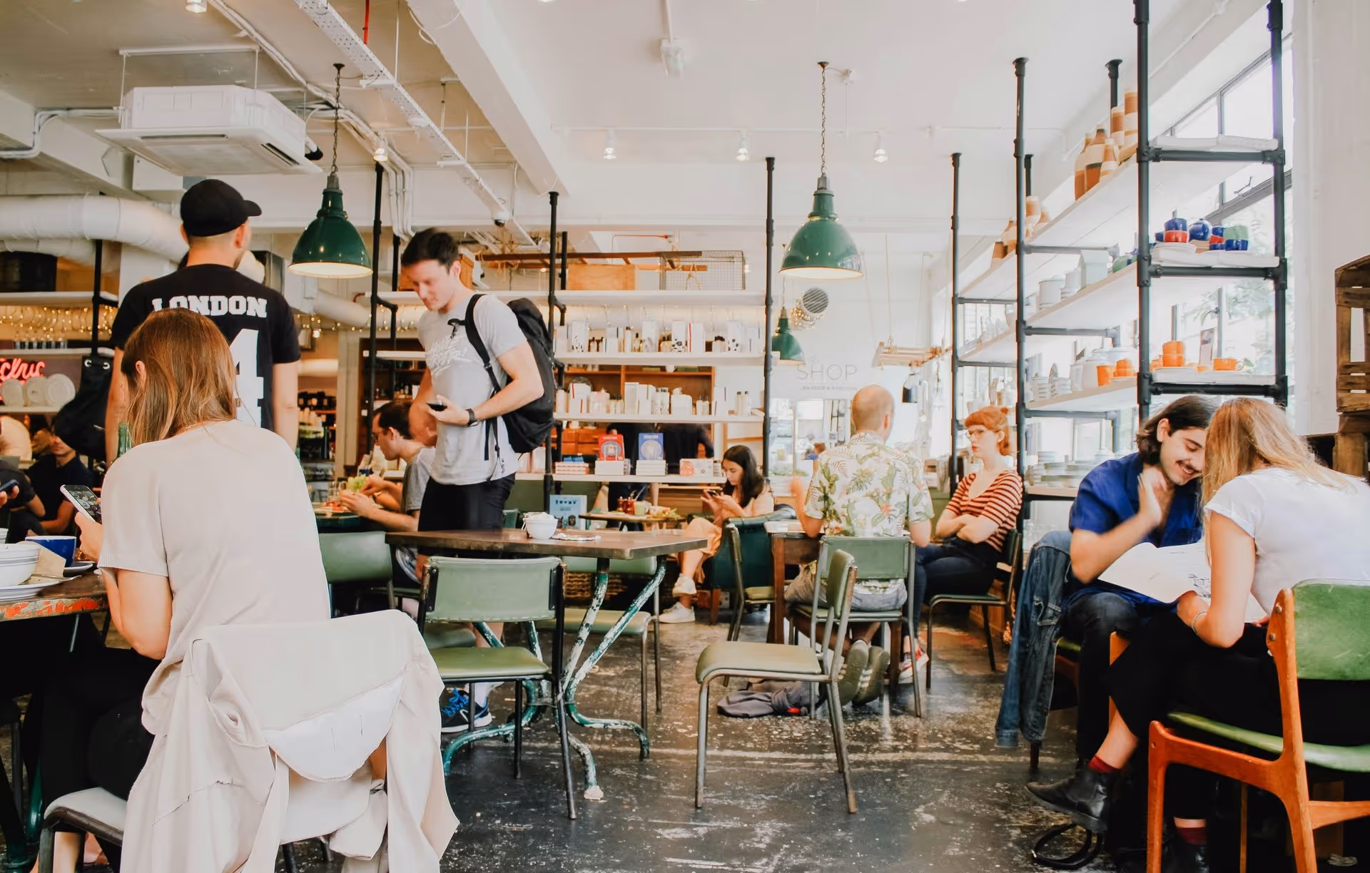 Busy cafe scene with people seated at tables, chatting and using phones. Bright lighting, modern decor. Warm, lively atmosphere.